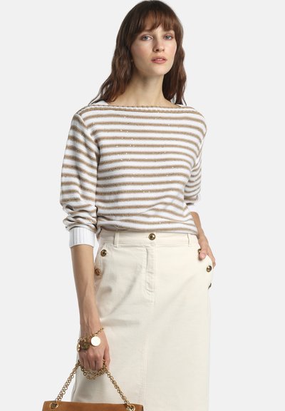Femme portant un pull beige et blanc à rayures et une jupe crème avec des boutons dorés, tenant un sac à main marron clair avec une bandoulière chaîne dorée.
