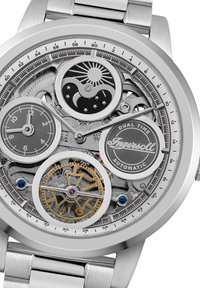 Montre automatique en argent dotée d'un cadran squelette complexe, de deux fuseaux horaires, et de rouages visibles avec des accents dorés et des pierres bleues.