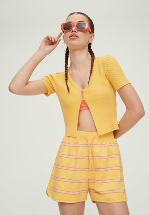 Cardigan court en maille côtelée jaune à manches courtes, associé à un short jaune avec des rayures roses. Le modèle porte des lunettes de soleil orange.