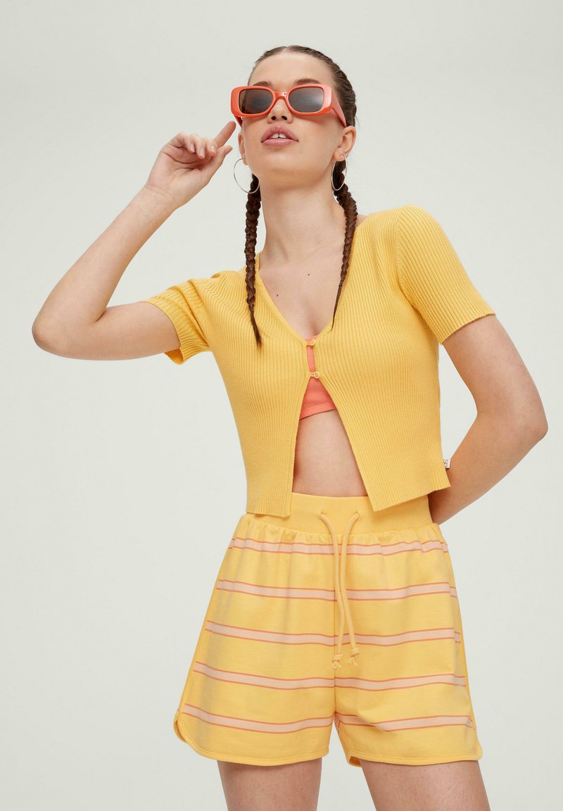Gelber gerippter Strick-Cropped-Cardigan mit kurzen Ärmeln, kombiniert mit gelben Shorts mit pinken Streifen. Das Model trägt orangefarbene Sonnenbrillen.