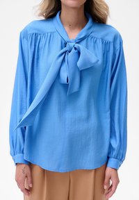 Kobieta w niebieskiej bluzce z długim rękawem z przewiązaną kokardą przy szyi i beżowych spodniach, stojąca z ramionami swobodnie opuszczonymi wzdłuż ciała.