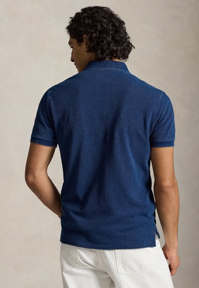 Man with dark curly hair wearing a navy blue short-sleeve polo shirt and white pants, standing turned away against a plain background.