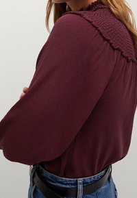 Bordeaux langmouw blouse met een smockhalslijn, zachte stof, ontspannen pasvorm en luchtige mouwen, gestyled met high-waisted denimjeans.