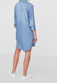 Light blue denim shirt dress with long sleeves, roll-tab cuffs, side slits, curved hem, and worn with white sneakers. Back view.