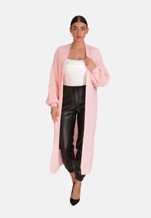 Woman wearing a long pink knit cardigan, white top, black leather jogger pants, black heels, and hoop earrings, posing against a white background.