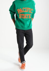 Person som bär en grön "PACIFIC STATE"-tröja, svarta jeans och orange sneakers, står med händerna bakom ryggen och ena benet över det andra.