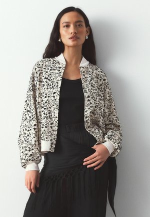 Woman wearing a cream floral bomber jacket over a black top and black skirt with fringe detail, standing against a plain white background.