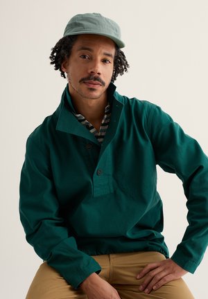 Muž kučeravých vlasov a s fúzmi, ktorý má na hlave svetlozelenú čiapku, na sebe tmavozelený pulóver a béžové nohavice, sedí na pozadí jednoduchej svetlej steny.