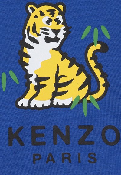Tigre aux rayures jaunes et noires assis parmi des feuilles vertes sur un fond bleu avec le texte "KENZO PARIS" en dessous.