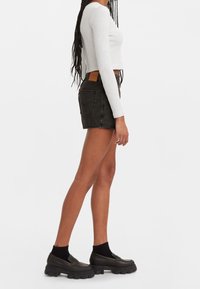 Kvinna med långt flätat hår iförd vit ribbad långärmad crop top, svarta denimshorts, svarta strumpor och svarta chunky loafers, stående på sidan.