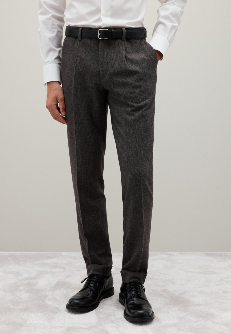 Pantaloni grigi su misura con un sottile motivo a quadri, fronte plissettato e una texture liscia. Abbinato a scarpe di pelle nere e una cintura nera.