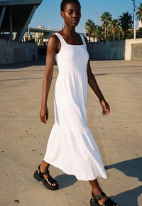Frau geht draußen spazieren, trägt ein ärmelloses weißes Maxikleid und schwarze Plateau-Sandalen an einem sonnigen Tag mit Palmen im Hintergrund.