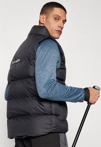 Black puffer vest with a high collar and horizontal stitching, paired with a textured blue long-sleeve shirt. Gray pants and a walking pole are included.