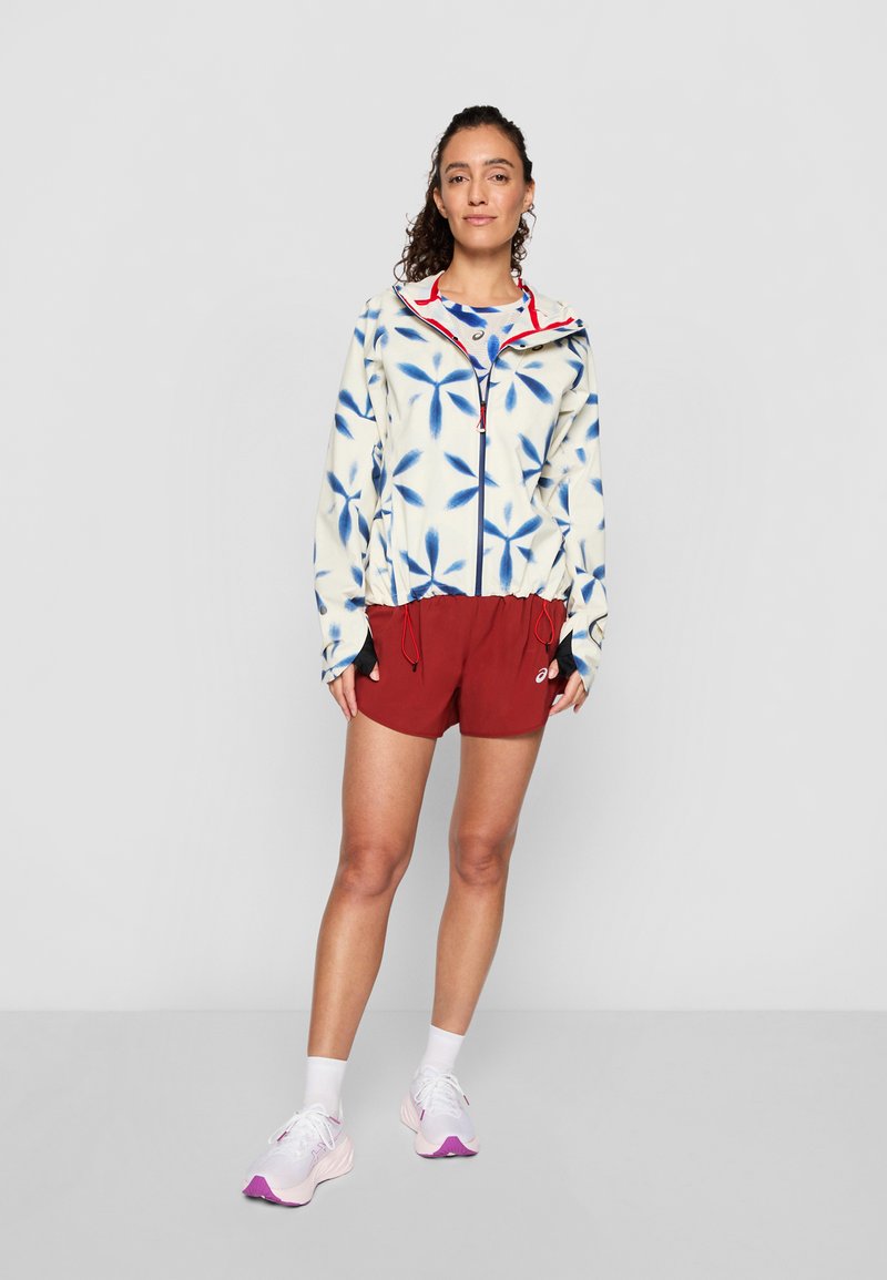 Junge Frau steht in einer weißen und blauen gemusterten Jacke, roten Sportshorts, weißen Socken und weißen Laufschuhen mit lila Sohlen.