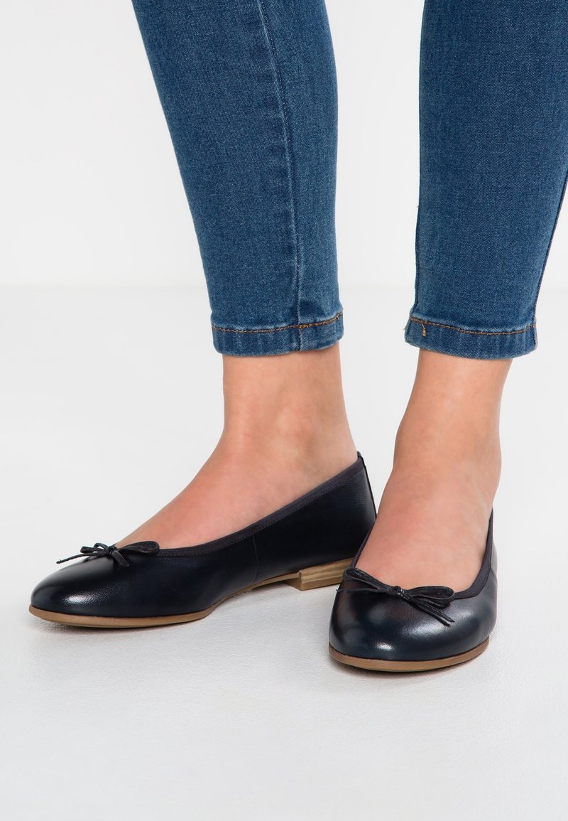 Pieds portant des ballerines en cuir bleu foncé avec de petits nœuds et des semelles marron clair, associées à un jean skinny bleu retroussé.