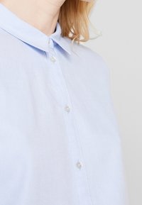 Hellblaues Hemd mit Button-Down-Kragen, aus strukturiertem Stoff und dünnen, weißen Knöpfen entlang der Vorderseite.
