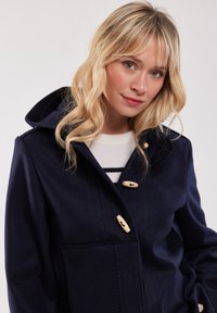Marineblå ullblandad kappa med huva, försedd med toggelknappar, fickor med lappar och texturerade sömmar. Enkla och funktionella design.