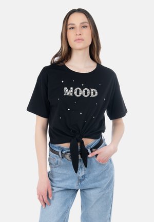 Frau trägt ein schwarzes, kurzärmeliges, geknotetes T-Shirt mit "MOOD" aus Strasssteinen, hellblaue Jeans und einen schwarzen Gürtel mit silberner Schnalle.
