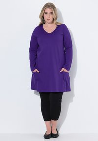 Lange lila Tunika mit V-Ausschnitt und zwei Fronttaschen, kombiniert mit schwarzen Leggings und flachen Schuhen, vor einem weißen Hintergrund.