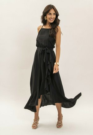 Femme souriante portant une robe noire sans manches avec un ourlet à volants, taille nouée, bracelet doré, boucles d'oreilles et sandales à talons hauts dorées à brides.