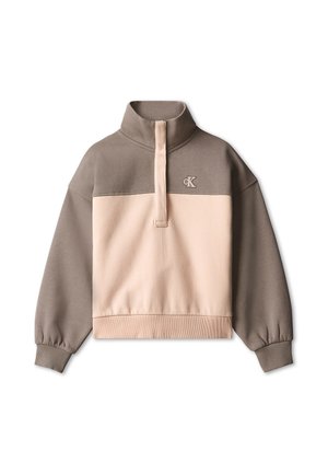Ung pige med brunt hår iført en pink og brun pullover, mørke jeans og sorte sko, står med benene spredte og smiler.