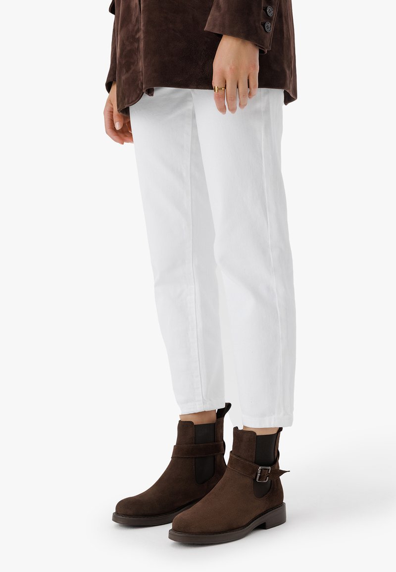 Brown suede ankle boots with a low block heel, elastic side panels, and an adjustable buckle accent, paired with white cropped jeans.