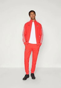 Rdeča trenirka iz tkanine z belimi črtami na rokavih in hlačah. Vključuje belo majico, zadrgo in podrobnosti z logotipom. Obuta je s črnimi športnimi copati.