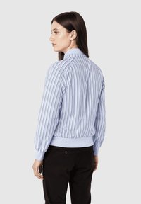 Mujer con cabello largo castaño, vestida con una blusa a rayas verticales en azul claro y blanco, y pantalones negros, de perfil frente a un fondo blanco.