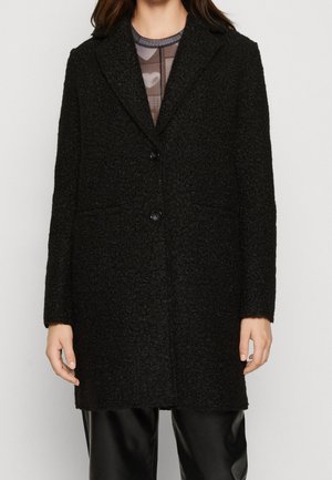 Femme portant un manteau en laine noire texturé avec deux boutons, un col et des manches longues, par-dessus un top transparent à motifs et un pantalon noir.