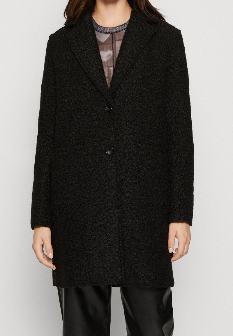 Femme portant un manteau en laine noire texturé avec deux boutons, un col et des manches longues, par-dessus un top transparent à motifs et un pantalon noir.