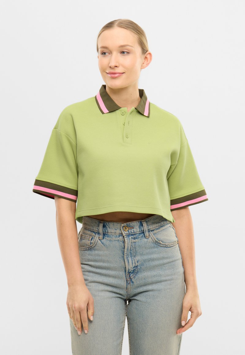 Nő világoszöld kivágott galléros pólóval, barna és rózsaszín csíkos gallérral és ujjakkal, világoskék magasderekú farmernadrággal párosítva.