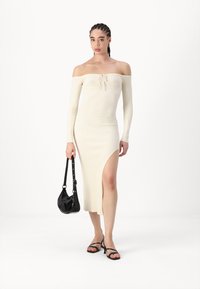 Robe beige côtelée à épaules dénudées avec manches longues, fente latérale et détail à cordon. Modèle tenant un sac à main noir, portant des sandales à lanières noires.