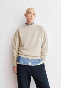 Beiges Sweatshirt mit rundem Ausschnitt und gesticktem Logo, darüber getragen ein blaues Jeanshemd mit lässiger Passform, kombiniert mit dunkler Hose.