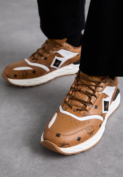 Brown leather sneakers with white accents, textured patterns, and gold hardware; thick white sole; round toe and lace-up design.