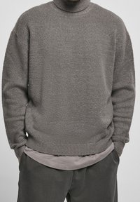 Maglione grigio a collo alto in maglia, con una texture morbida, vestibilità rilassata, e polsini e orlo a coste. Indossato sopra una camicia leggera grigia a strati.