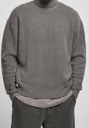 Pull à col roulé en tricot gris avec une texture douce, une coupe décontractée, et des poignets et un ourlet côtelés. Porté par-dessus une chemise superposée gris clair.