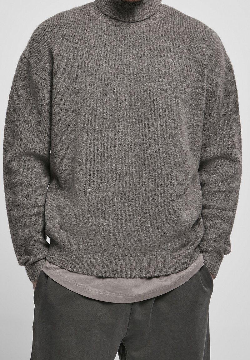 Maglione grigio a collo alto in maglia, con una texture morbida, vestibilità rilassata, e polsini e orlo a coste. Indossato sopra una camicia leggera grigia a strati.