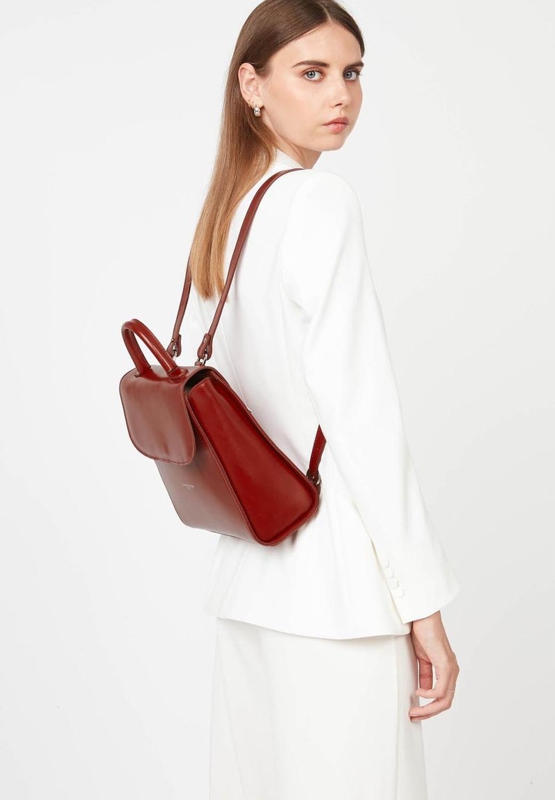 Sac à dos en cuir rouge avec un design structuré, comportant une poignée supérieure et des bretelles réglables. Texture lisse et détail de fermeture à rabat.