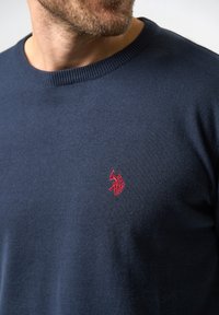 Mørkeblå sweater lavet af et glat strikket stof. Har en rund hals og et rødt broderet logo på venstre bryst.