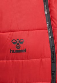 Tissu matelassé rouge avec logo "hummel" noir et symbole d'abeille à côté d'une bande verticale noire présentant le texte "hummel" répété.