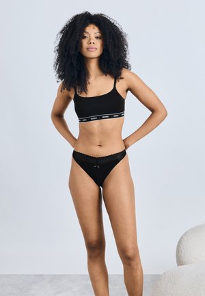 Zwarte bralette met dunne bandjes en een logoband; bijpassende zwarte string met kantafwerking en strikdetail. Soepel, rekbaar materiaal.