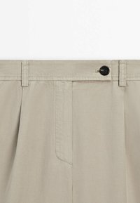 Beige bukser med flad front, to folder og en enkelt knaplukning. Har sidelommer og en glat, vævet tekstur.