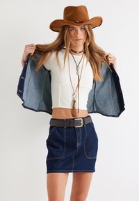 Chaqueta de mezclilla con interior en lavado claro, top corto blanco ajustado y falda de mezclilla oscura. Un sombrero de vaquero de fieltro marrón y un collar en capas complementan el look.
