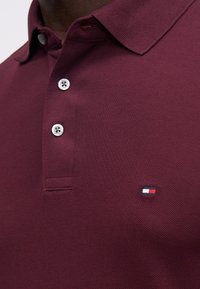Polo bordeaux em tecido texturizado, com uma pala de dois botões e um pequeno logótipo bordado em azul, branco e vermelho.