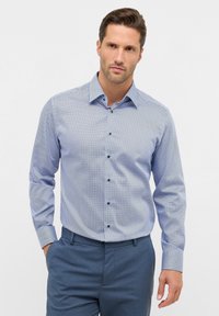 Blauwe overhemd met knopen met een klein geometrisch patroon, lange mouwen en een kraag. De stof heeft een gladde textuur. Gecombineerd met blauwe broeken.