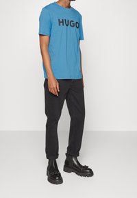 Niebieska bawełniana koszulka z wyraźnym czarnym logo "HUGO", zestawiona z czarnymi zwężającymi się spodniami i czarnymi botkami Chelsea z teksturowanymi akcentami.