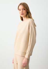 Felpa beige a girocollo con maniche lunghe e dalla vestibilità ampia. Presenta una striscia testurizzata lungo la cucitura della spalla. Tessuto morbido con una finitura liscia.