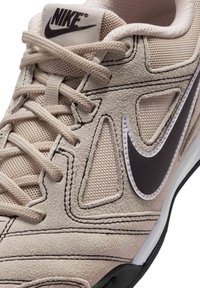 Μπεζ αθλητικό παπούτσι Nike με λεπτομέρειες από δίχτυ και σουέντ, μαύρο λογότυπο Nike, επενδεδυμένη γλώσσα και μπεζ κορδόνια.