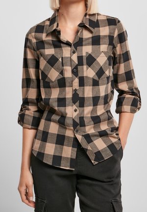 Woman wearing beige and black checkered button-up shirt with rolled-up sleeves and black pants with hands partially in pockets.
