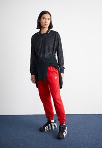 Camisa preta de mangas longas com textura geométrica, combinada com calças de treino vermelhas brilhantes. Ténis em preto e branco com um design arrojado. Suéter de lã amarrado na cintura.